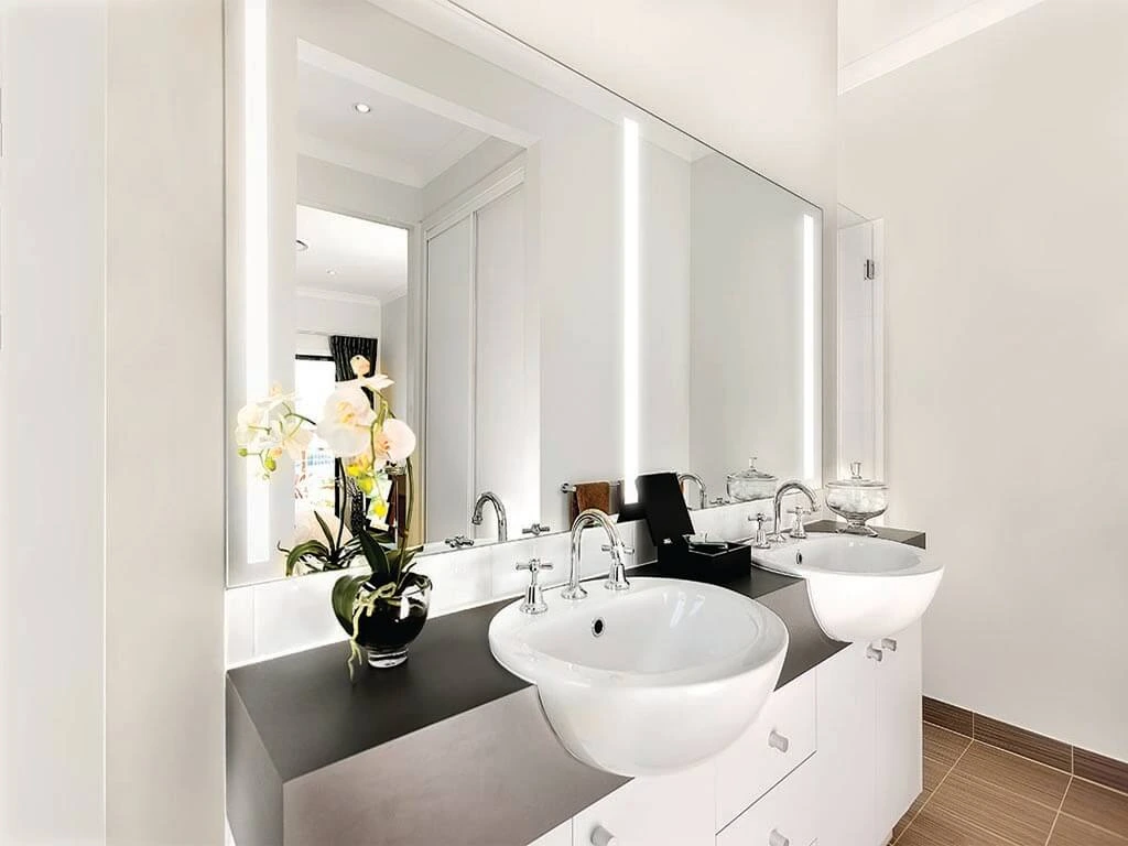 A double sink lighted mirror on a white wall with a flower on a black vase under the cabinet sink.